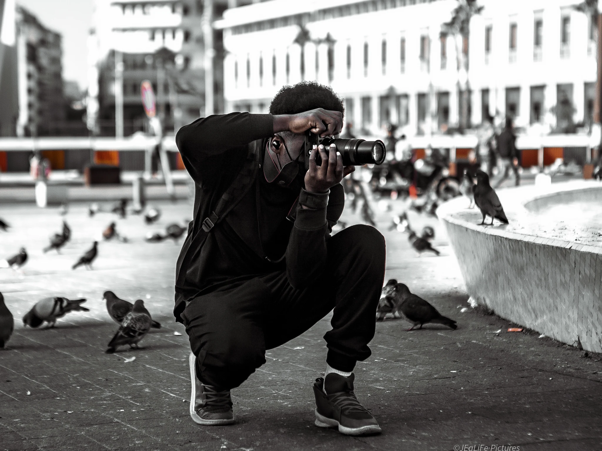 Jean Guylane Memiaghe Biteghe accroupi, appareil photo à l'œil, au milieu des pigeons